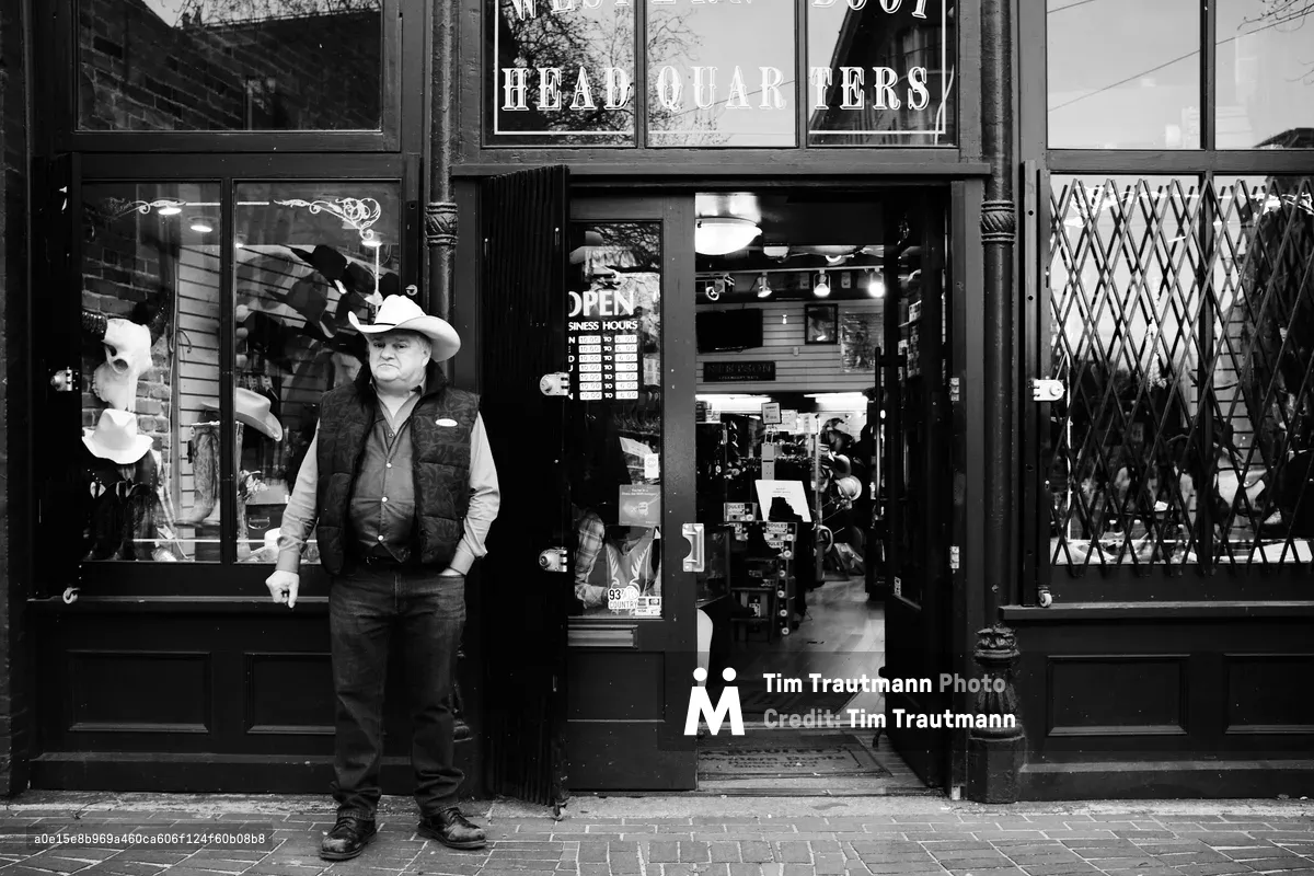 Tony Chisholm, a sales associate at OK Boot Corral, takes a contemplative break outside the Western wear store in Vancouver's historic Gastown district. Dressed in authentic cowboy attire—white Stetson hat, dark vest, and weathered boots—he leans casually against the ornate storefront of Byrnes Block on Carrall Street. The black and white composition captures the timeless intersection of frontier heritage and urban commerce, with vintage display windows revealing leather goods and Western memorabilia within.