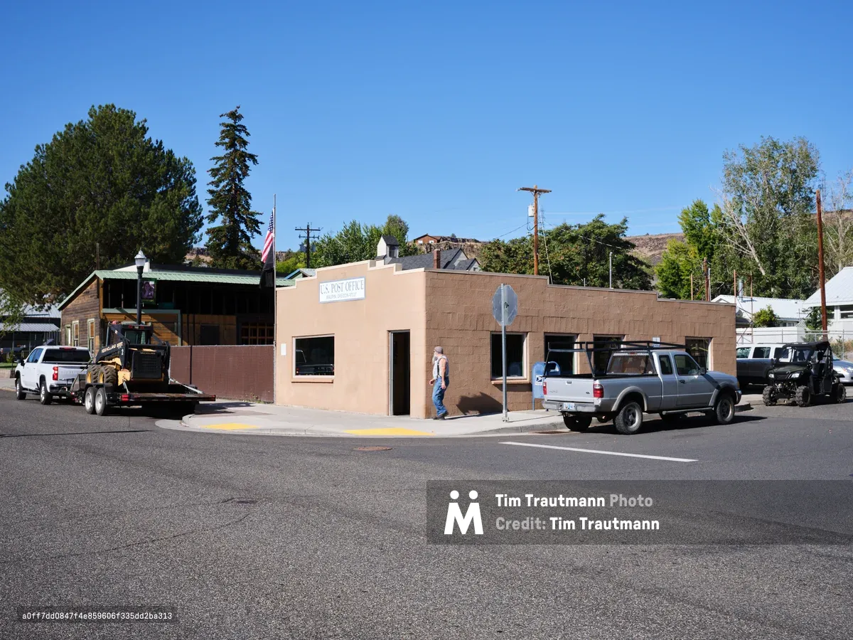 A modest brick post office stands as the communication lifeline for Maupin, Oregon, its weathered facade catching the high desert sun beneath an endless azure sky. Pickup trucks line the street while a figure in work clothes approaches the entrance, embodying the quiet rhythm of rural American life. The Stars and Stripes flutter above neighboring buildings, while tall pines and distant hills frame this essential community cornerstone in the Columbia River Gorge region.