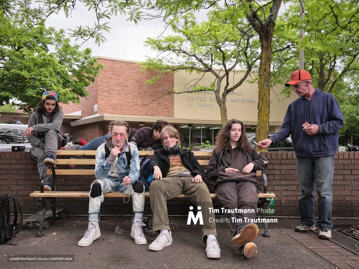 A group of five teenagers hang out on a public bench in the town square of Portland's St. Johns neighborhood. The teens wear a mix of hoodies, ripped jeans, and white sneakers, with skateboards visible beside them. One wears pink-tinted sunglasses, another has headphones around his neck. An older man in an orange Nike cap and blue hoodie stands to the right, offering two cigarettes to one of the teenagers. A brick United States National Bank of Oregon branch building is visible in the background, framed by leafy green trees under an overcast sky.