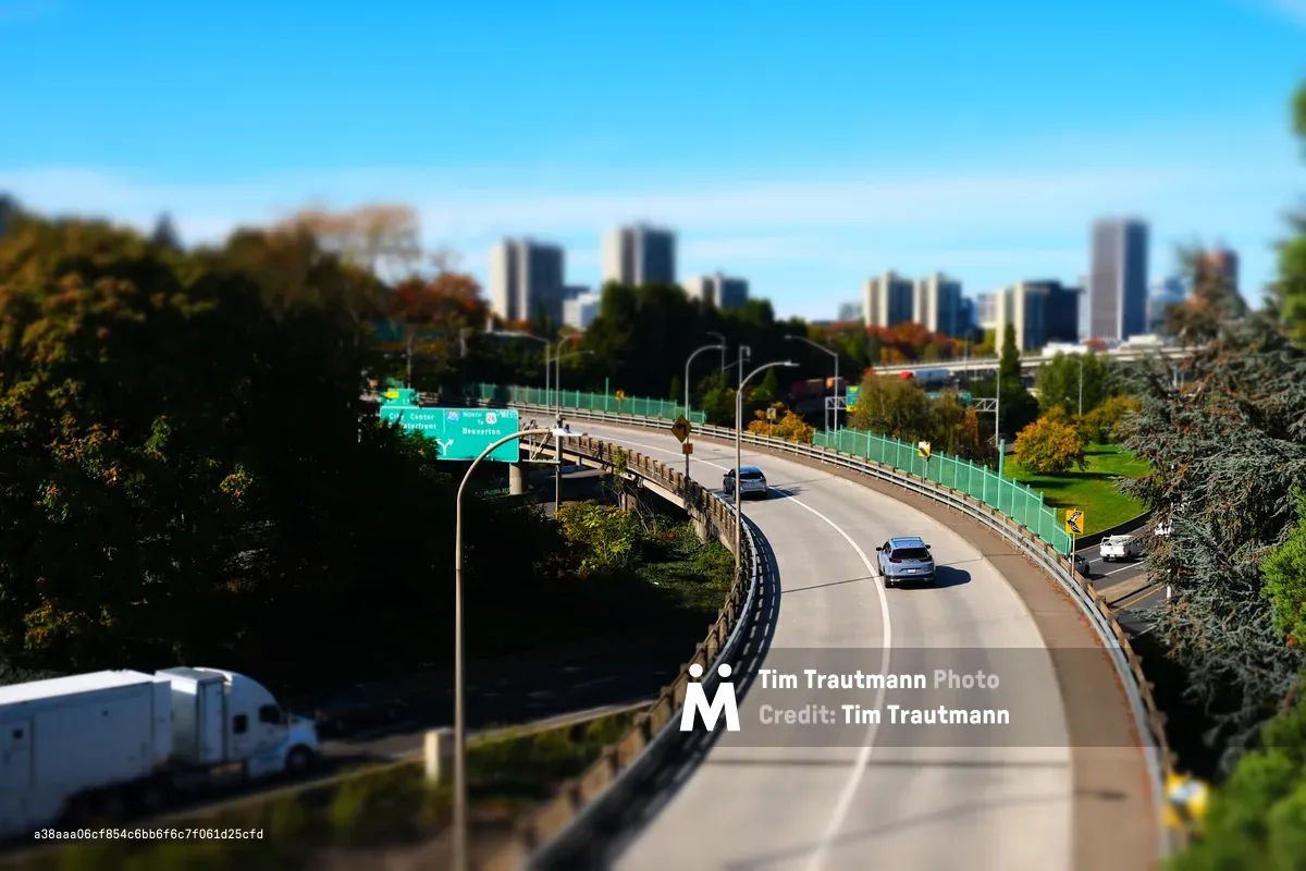 A tilt-shift photograph of a curving overpass ramp in South Portland, Oregon, giving the scene a miniature model-like appearance. Two cars navigate the sweeping curve toward downtown, while a semi-truck is visible on a lower road to the left. Green highway signs point toward City Center, Waterfront, and Beaverton. Autumn trees in orange, yellow, and green frame both sides of the road, and the Portland downtown skyline rises in the soft-focus background under a vivid blue sky.