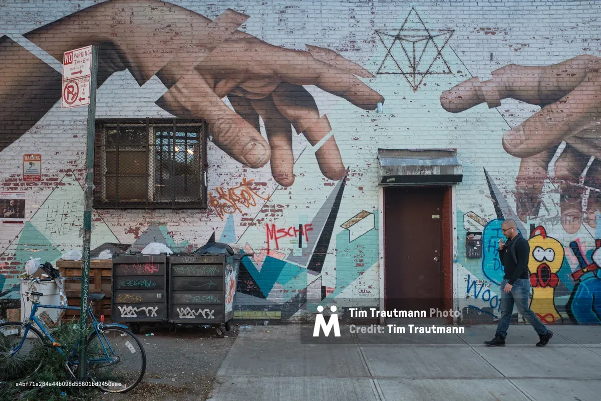 A lone figure in a black leather jacket strides past a vibrant mural on weathered brick walls in Williamsburg, Brooklyn. The street art features massive reaching hands, geometric patterns, and colorful character illustrations that transform the urban facade into a canvas of contemporary expression. A blue bicycle rests against dumpsters in the foreground, while peeling paint and layered graffiti tags speak to the neighborhood's authentic street culture. The overcast lighting creates a moody atmosphere that captures the raw creative energy of Brooklyn's artistic landscape.