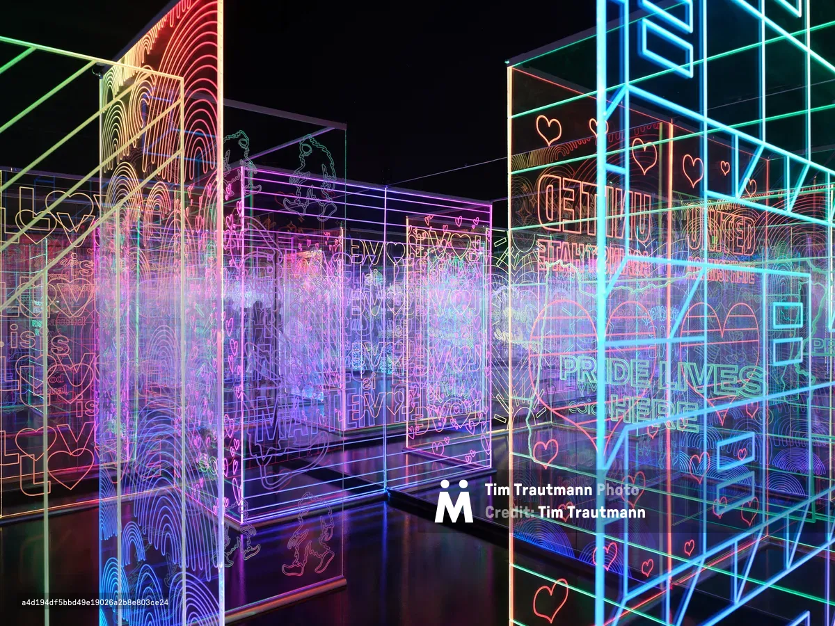 A vibrant interactive art installation featuring glowing neon geometric shapes, wireframe structures, and heart symbols in blues, pinks, and greens displayed in a dark gallery space. The luminous digital art creates an immersive environment with overlapping transparent panels and geometric forms.