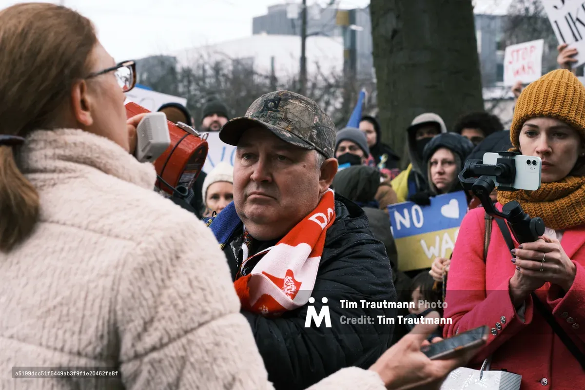 In the gray winter light outside Portland's Revolution Hall, tension crackles through a crowd gathered in the days before Russia's invasion of Ukraine. A middle-aged man in a camouflage cap stands resolute amid the pressing crowd, his weathered face betraying quiet determination as protesters surge around him with signs and smartphones raised. The intimate proximity of opposing voices creates a palpable friction, while bare trees and urban architecture frame this moment of civic discord on Southeast Stark Street.