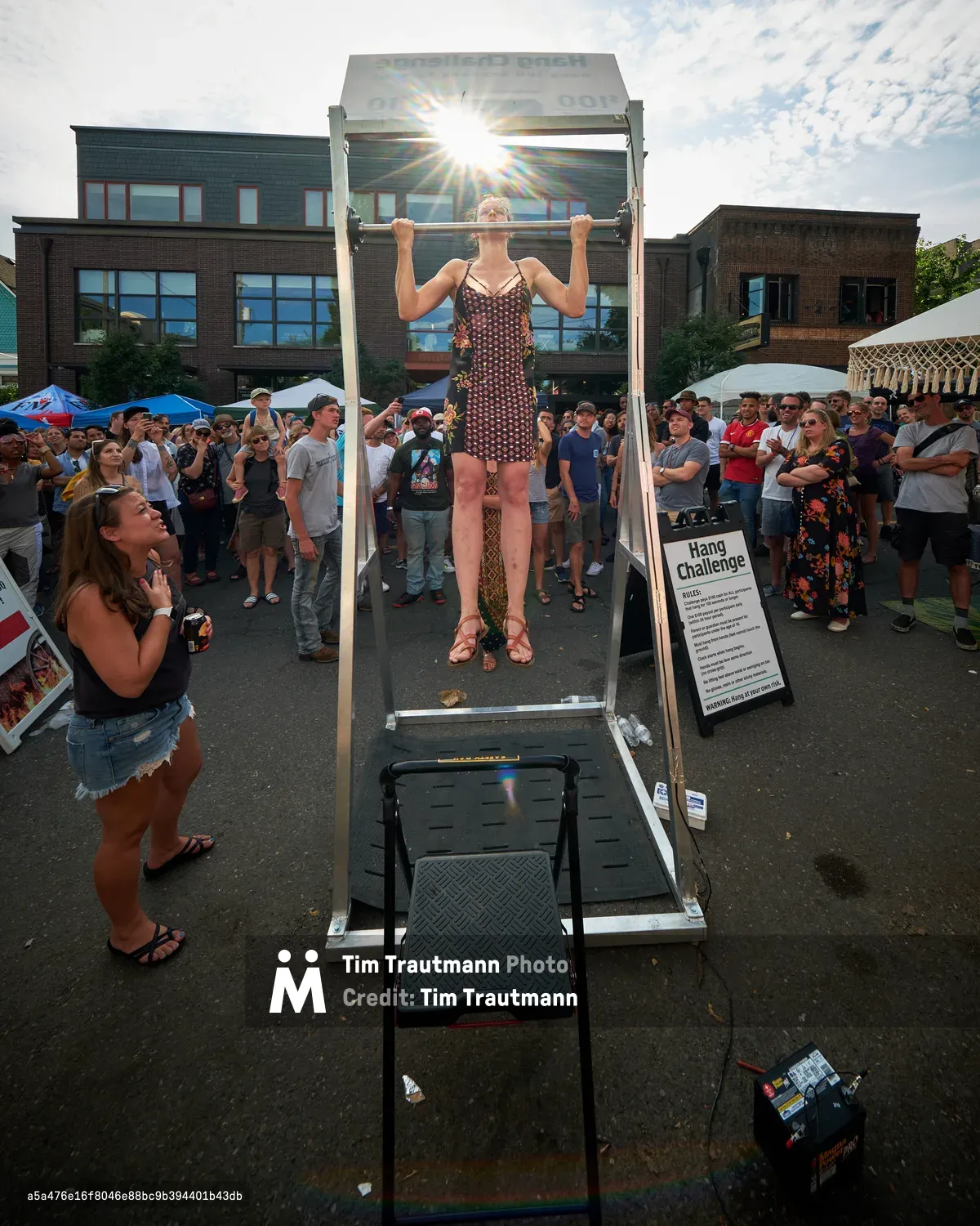A determined woman in a geometric patterned dress executes a perfect dead hang from a pull-up bar at Portland's Mississippi Street Fair, her form silhouetted against the brilliant afternoon sun. The crowd of onlookers forms a semicircle around the Hang Challenge station, their faces a mixture of encouragement and awe as she demonstrates raw strength in an unexpected urban setting. The lens flare creates dramatic rays that pierce through the scene, while the contemporary brick architecture of North Mississippi Avenue provides a distinctly Portland backdrop to this moment of athletic determination.