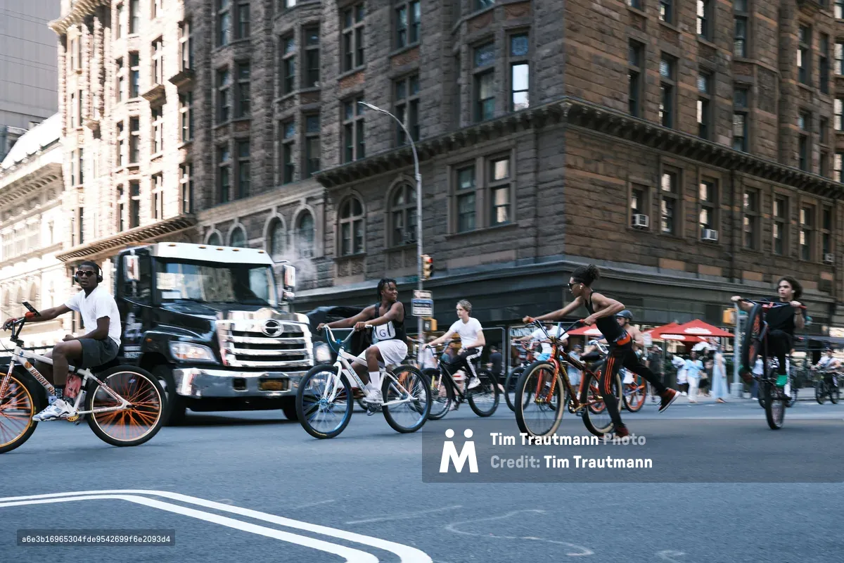 A dynamic street scene captures cyclists weaving through traffic on 7th Avenue in Manhattan, their silhouettes moving against the weathered stone facades of historic buildings. The golden hour light bathes the pre-war architecture in warm tones while creating dramatic shadows across the asphalt. A diverse group of riders navigates the urban landscape alongside a delivery truck, embodying the rhythm and energy of New York City street life.