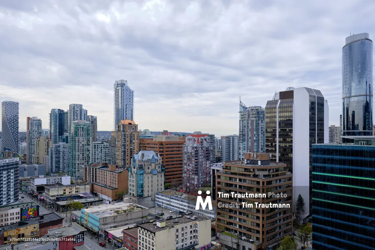 A sweeping aerial view of Vancouver's dense downtown core captured beneath a dramatic canopy of pewter clouds. The urban landscape unfolds in layers of architectural diversity, from gleaming glass towers to weathered brick facades, creating a complex metropolitan mosaic. Muted daylight filters through the overcast sky, casting the city in subdued tones that emphasize the geometric interplay between old and new structures. The composition reveals the organic density of urban development, where residential high-rises, commercial buildings, and street-level establishments converge in Vancouver's characteristic west coast urbanism.