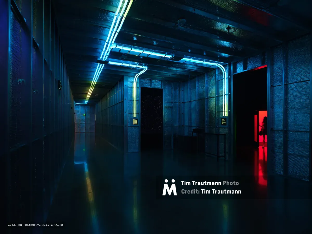 A dramatic underground corridor featuring flooded concrete floors that reflect vibrant blue and red neon lighting from overhead fixtures. The industrial space has exposed pipes and ventilation systems along the ceiling, creating a cyberpunk aesthetic with strong architectural lines leading into darkness.