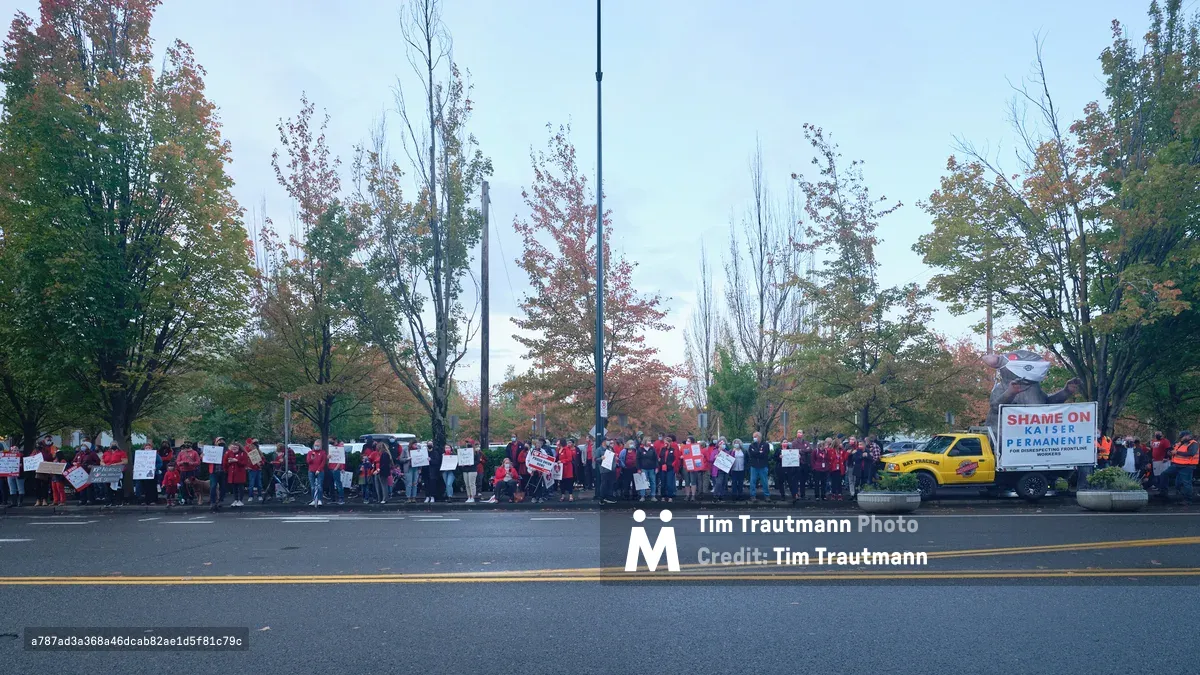 A sea of red-clad nurses and supporters mass along Northeast Multnomah Street outside Kaiser Permanente Tower in Portland's Lloyd District, their protest signs creating a rhythmic pattern against autumn's burnished foliage. The overcast Pacific Northwest sky casts diffused light across the demonstration, while a yellow union truck bearing "SHAME ON KAISER PERMANENTE" anchors the right side of the frame. The striking healthcare workers stretch in an unbroken line along the sidewalk, their collective presence transforming the corporate streetscape into a theater of labor solidarity.