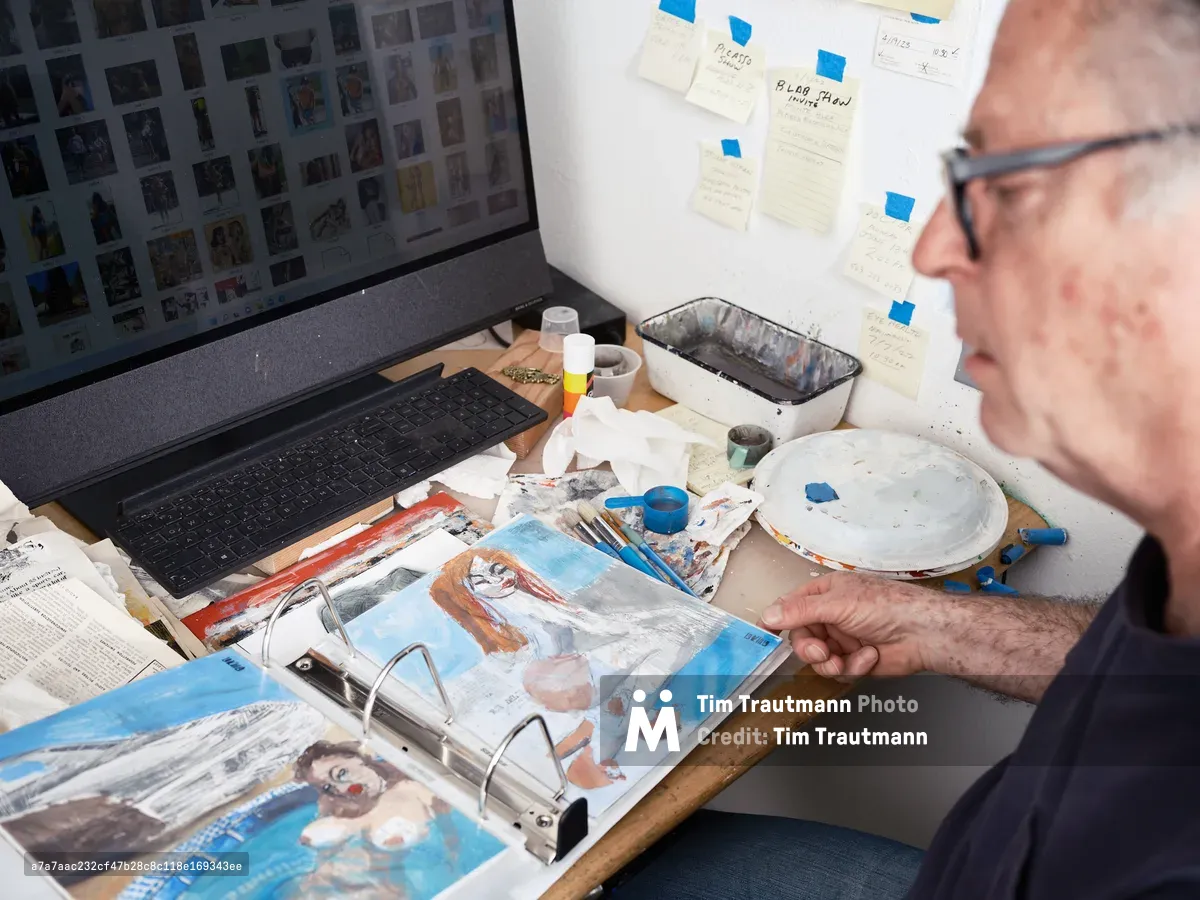 An older man with glasses works on colorful paintings at a cluttered desk in his art studio, with a laptop displaying reference images and art supplies scattered around. The workspace shows an active creative process with paint palettes, brushes, and artwork in various stages of completion.