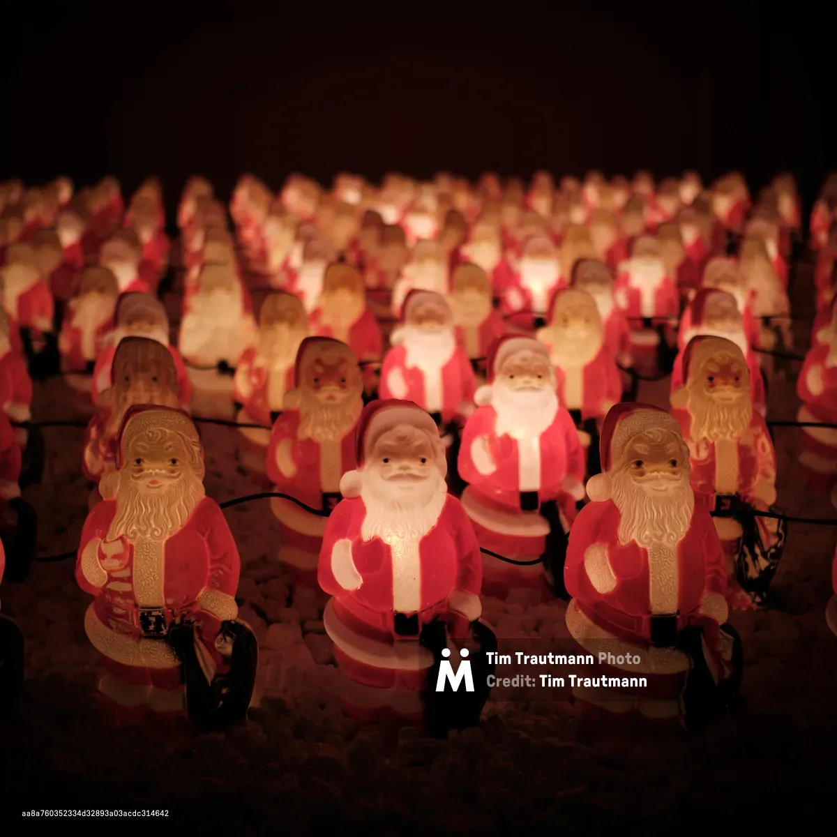 An enchanting nocturnal scene unfolds in Portland's Concordia neighborhood where hundreds of illuminated Santa Claus figurines create a surreal winter wonderland. The glowing plastic Santas, arranged in perfect rows, emanate a warm amber light that pierces the velvety darkness, their jolly faces creating an almost hypnotic pattern that stretches into the shadows. Chris Willis's annual Santa Clones installation transforms this Northeast Portland corner into a whimsical spectacle, where mass-produced holiday cheer takes on an unexpectedly artistic quality. The soft bokeh effect in the background dissolves the distant figures into dreamlike orbs of red and gold, creating depth and mystery in this delightfully surreal Christmas tableau.