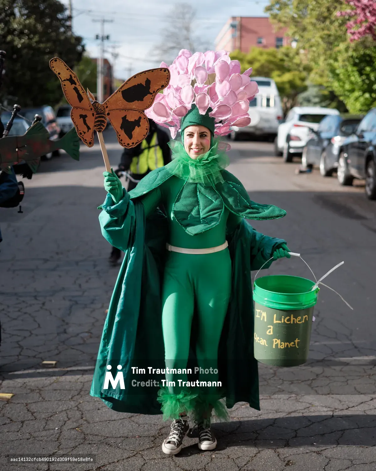 A person dressed in an elaborate green plant costume with pink flowers stands on a cobblestone street in Portland, Oregon, holding a butterfly prop and a bucket with environmental messaging. The costume includes leafy details and the bucket reads about creating a clean planet.