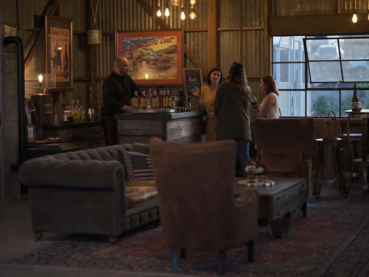 The warmly lit interior of the cocktail bar at Abbey Road Farm in Carlton, Oregon. A bartender in a black shirt stands behind a rustic wooden bar stocked with bottles of whiskey, as three women chat and enjoy drinks. Vintage whiskey advertising posters, including ones for Uam Var Scotch Whisky and Dunard Dew Old Irish Whiskey, hang on corrugated metal walls. Edison bulbs dangle from the ceiling, and the eclectic lounge area in the foreground features tufted velvet sofas, worn leather armchairs, and a Persian rug. A large industrial window lets in soft evening light on the right.