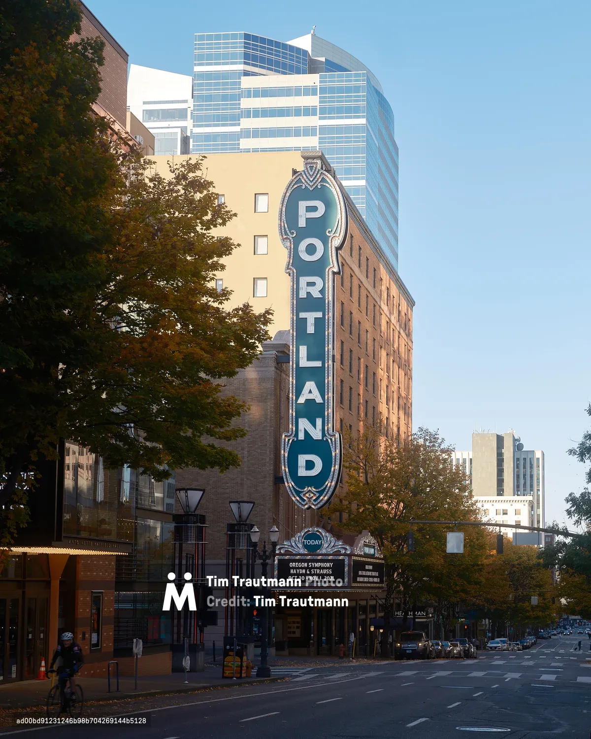 The exterior of the Arlene Schnitzer Concert Hall on Broadway in downtown Portland, Oregon, photographed on a clear autumn day. The venue's iconic large teal and white vertical "Portland" marquee sign dominates the facade of the historic brick building. The theater's marquee below reads "Oregon Symphony — Haydn & Strauss — Oct 30 - Nov 1, 2021." Autumn trees in golden and orange foliage frame the street, and a cyclist rides past in the foreground. The Heathman Hotel is visible further down the block, with modern glass office towers rising behind the historic building against a clear blue sky.