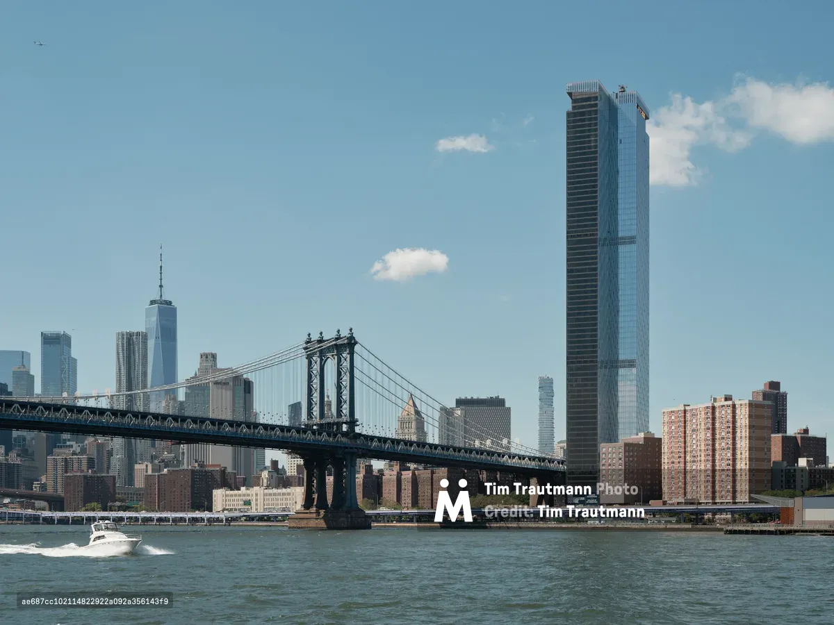 The iconic Manhattan Bridge stretches across the East River in elegant suspension, its steel towers and cables creating geometric lines against a pale blue afternoon sky dotted with cotton-white clouds. One World Trade Center's gleaming spire punctuates the left side of Manhattan's financial district, while a striking dark glass tower dominates the right portion of the frame. A white pleasure boat cuts through the choppy river waters in the foreground, leaving a gentle wake as it passes beneath this architectural symphony of old and new New York.