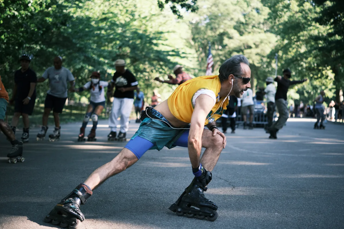 A man in a vibrant yellow tank top and teal shorts leans into a graceful skating stride along Central Park's sun-dappled pathways. The afternoon light filters through the dense canopy above, creating a mosaic of shadows on the asphalt as other skaters and park visitors blur into the verdant background. His focused expression and fluid movement capture the meditative rhythm of urban recreation, while the American flag glimpsed among the trees anchors this quintessentially New York moment.