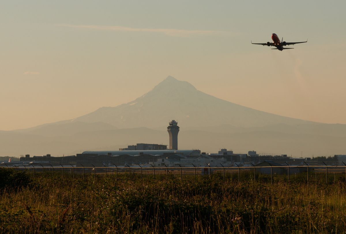 A commercial aircraft flies through a hazy sky above an airport control tower and terminal buildings, with a large mountain silhouetted in the background during golden hour lighting. The foreground shows wild grass and vegetation with a security fence marking the airport perimeter.