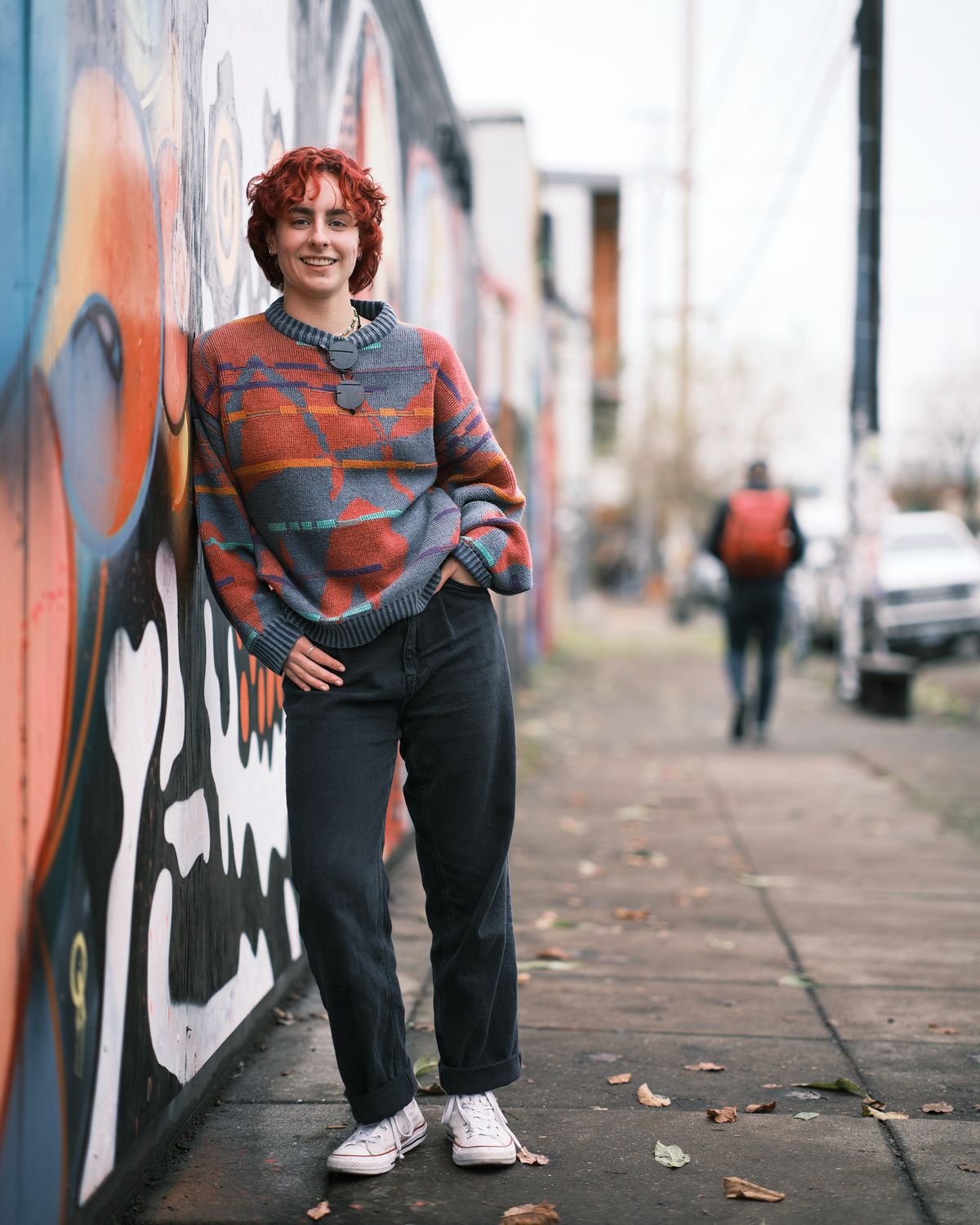 A full-length senior portrait of Annika Molina leaning against a colorful street mural on a Portland sidewalk. She has short curly red hair and wears an oversized patterned sweater in orange, grey, and teal, dark wide-leg jeans cuffed at the ankle, white Converse sneakers, and a pendant necklace. She smiles warmly at the camera with one hand on her hip. A pedestrian with a red backpack walks in the blurred background along the tree-lined street.