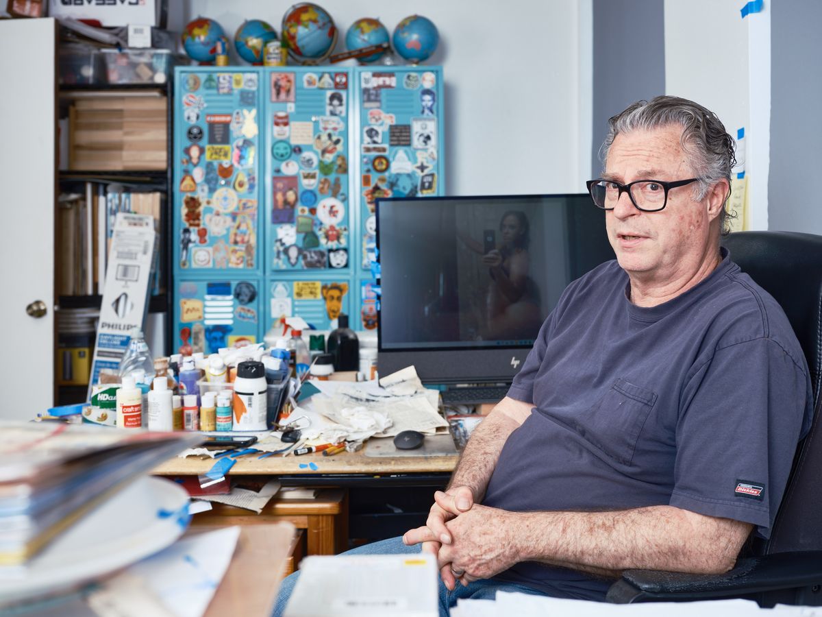 A middle-aged man with glasses sits in his cluttered creative studio workspace, surrounded by art supplies, globes, and colorful displays on the walls. The space shows an active artist's environment with paints, papers, and various creative materials scattered across his desk.