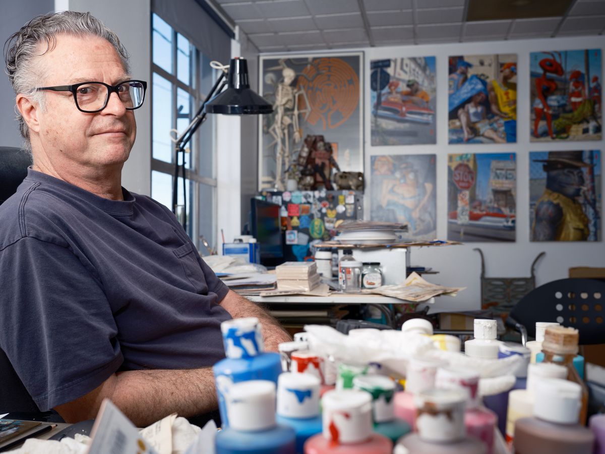 A middle-aged man with glasses and gray hair sits in his art studio surrounded by colorful paint tubes, brushes, and artwork displayed on the walls. The bright, organized workspace features large windows, a desk lamp, and various artistic materials scattered throughout the creative environment.
