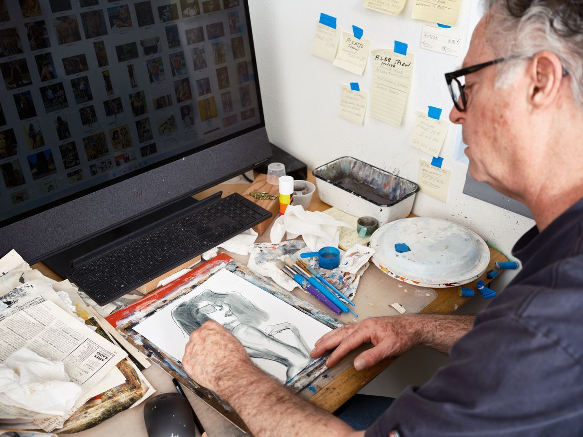 An older man with glasses sits at a cluttered desk in his Portland art studio, working on a portrait drawing while surrounded by art supplies, reference images on his laptop screen, and notes taped to the wall.