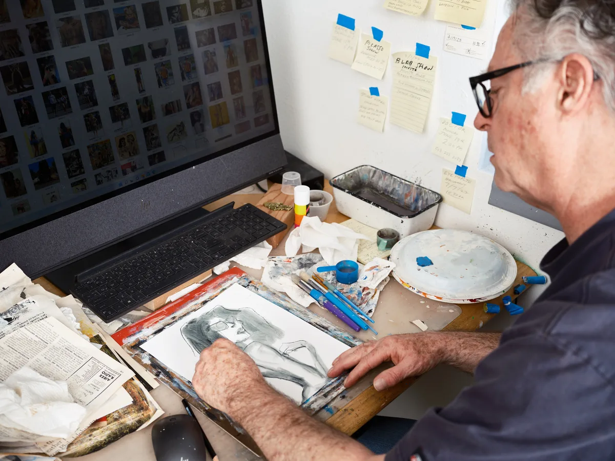 An older man with glasses sits at a cluttered desk in his Portland art studio, working on a portrait drawing while surrounded by art supplies, reference images on his laptop screen, and notes taped to the wall.