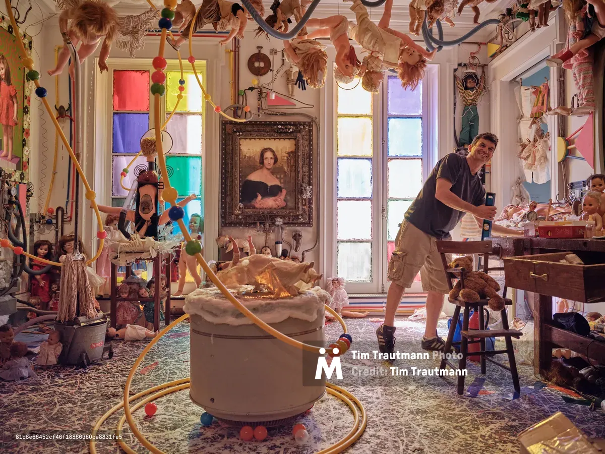 Artist Mikel Glass navigates through his fantastical installation space at The Cell Theatre on West 23rd Street, surrounded by an explosion of suspended dolls, colorful geometric window panels, and eclectic found objects. The scene pulses with creative energy as Glass moves through this immersive wonderland of theatrical props and artistic debris. Natural light filters through the rainbow-hued window panes, casting jewel-toned shadows across the cluttered floor where vintage portraits and whimsical toys create a dreamlike tableau. The atmosphere captures the raw, unfiltered moment before an artistic transformation unfolds.