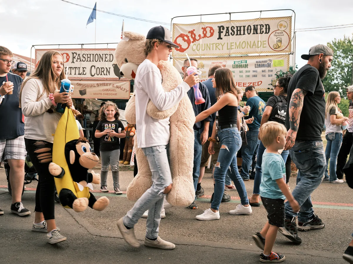 A bustling crowd of fairgoers walks through the Clark County Fair in Vancouver, Washington. In the center of the frame, a teenage boy in a white sweatshirt and black cap carries an enormous stuffed teddy bear he won at a carnival game. To his left, a girl carries a large stuffed monkey in a banana costume. Families, children, and adults fill the midway, with food vendor signs for Old Fashioned Country Lemonade and Old Fashioned Kettle Corn visible in the background. Several people carry colorful fair drinks.