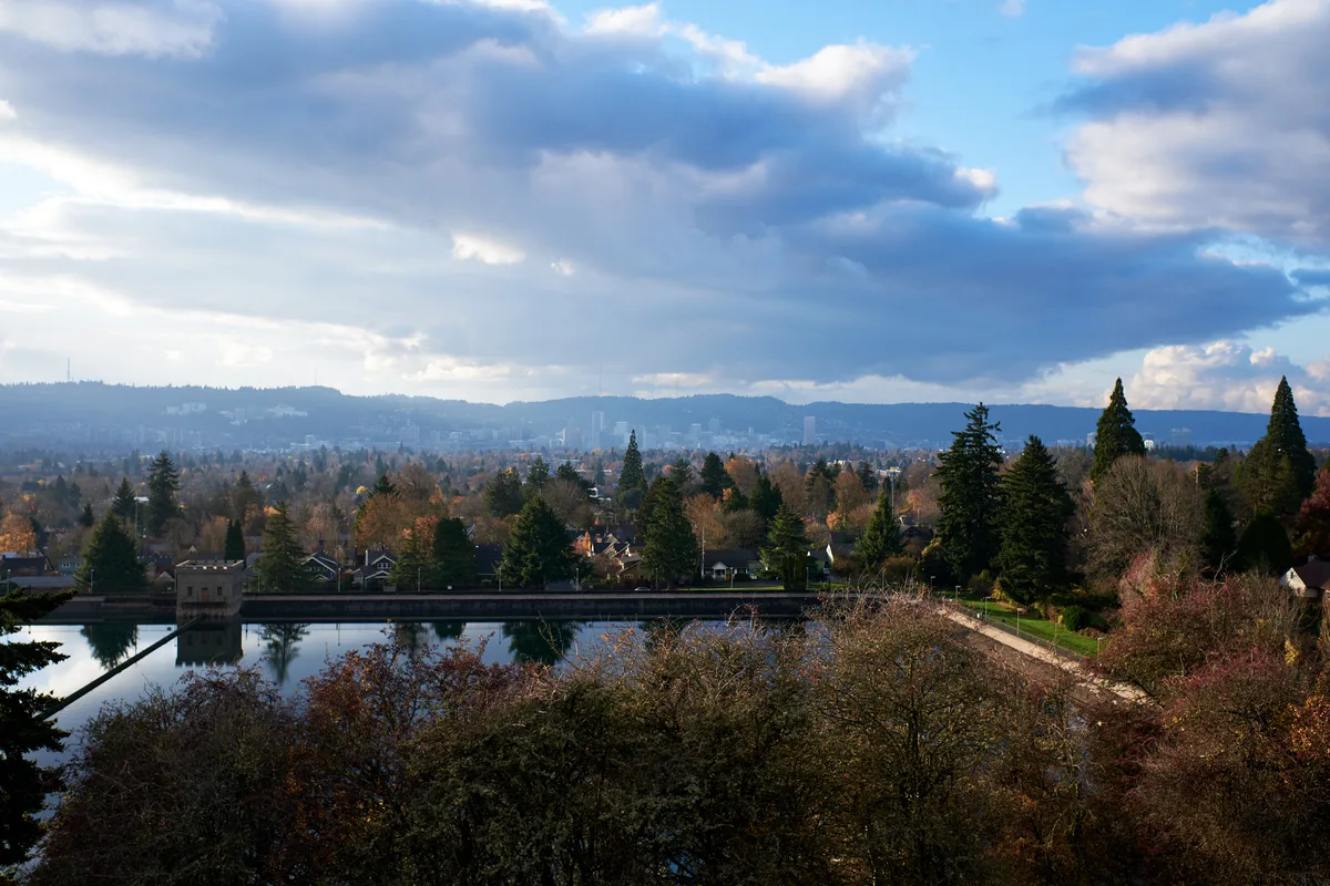 Storm clouds gather dramatically over Portland's residential sprawl as viewed from Mount Tabor's reservoir overlook during peak autumn foliage. The serene water surface mirrors the moody sky while evergreen sentinels frame the foreground, their dark forms contrasting against the warm amber and rust tones of deciduous trees below. The city extends toward distant hills under brooding cumulus formations, creating a contemplative tableau that captures the Pacific Northwest's characteristic interplay between urban development and natural beauty.