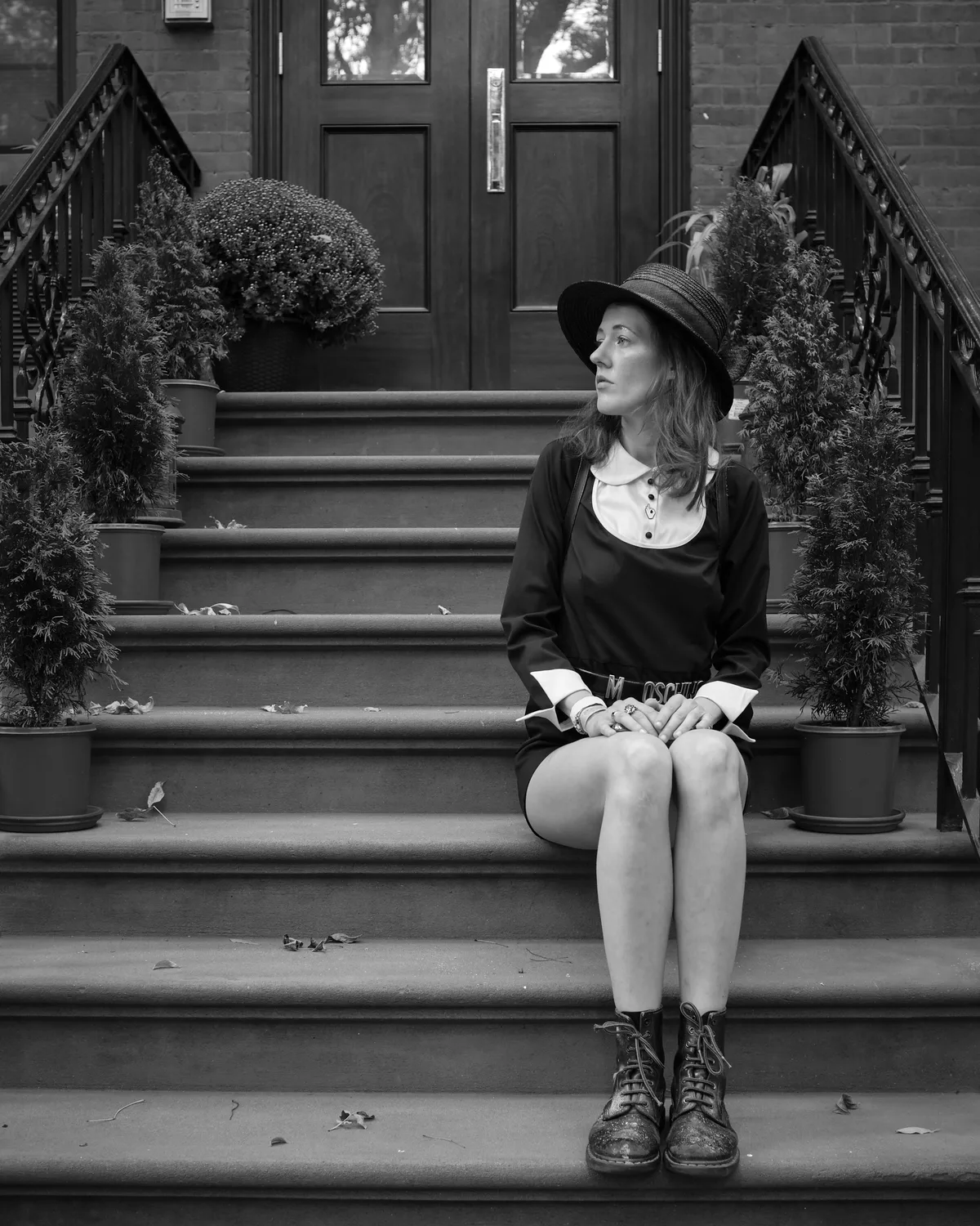 A young woman in vintage-inspired attire sits contemplatively on weathered stone steps, her gaze cast thoughtfully to the side beneath the shadow of a wide-brimmed hat. The monochromatic composition captures the intimate solitude of urban autumn, with ornate iron railings framing seasonal plantings that speak to Brooklyn's historic brownstone character. Fallen leaves scattered across the steps and her classic lace-up boots ground the scene in transitional seasonal beauty.