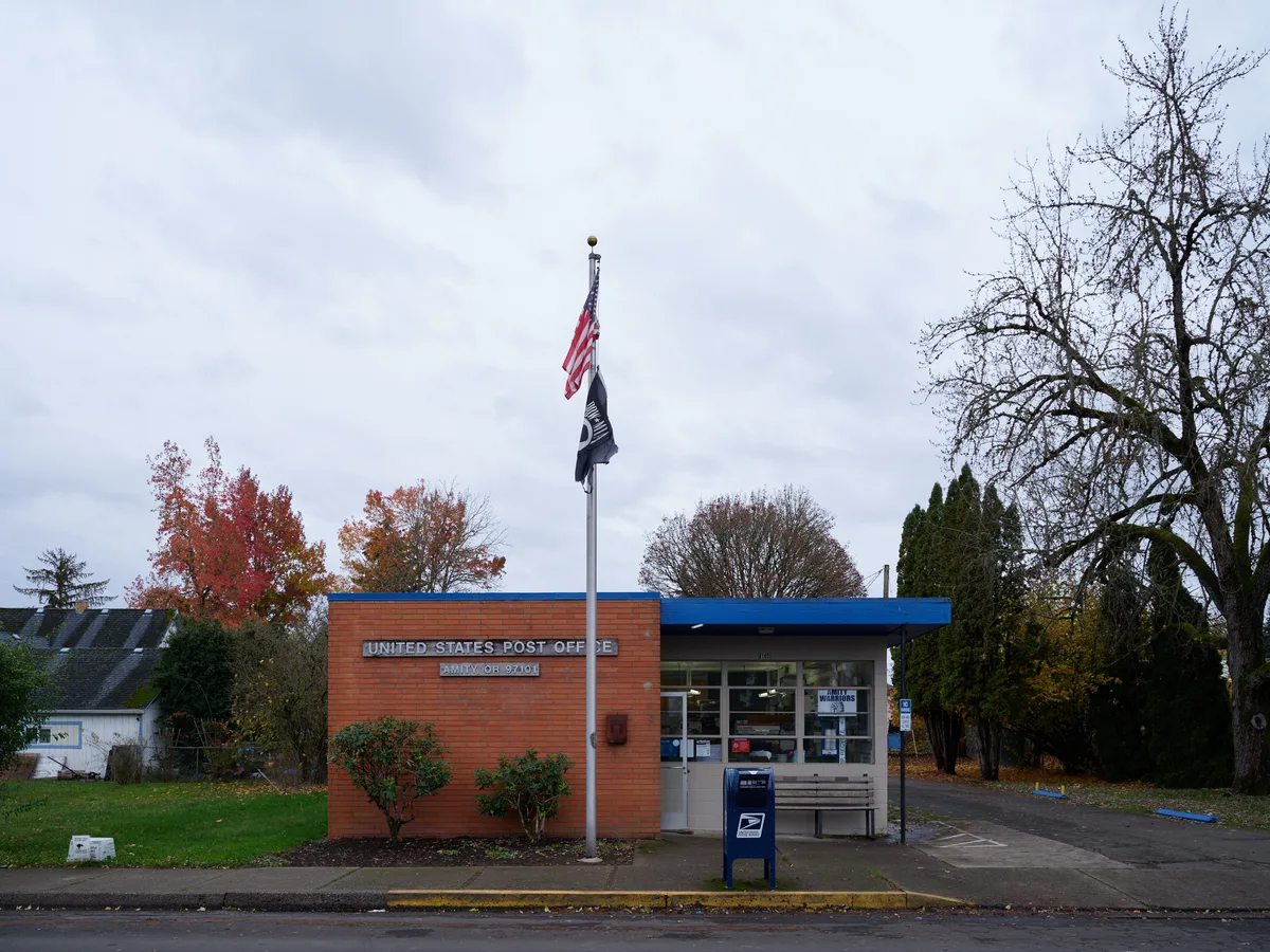 Under a pewter November sky, the modest brick facade of Amity's post office stands as a cornerstone of rural community life. The building's warm terracotta walls contrast with its bright blue trim, while overhead flags flutter in the crisp air. Surrounded by deciduous trees displaying their final burst of scarlet and gold, this quintessential small-town postal station embodies the enduring presence of federal service in Oregon's Yamhill County countryside.