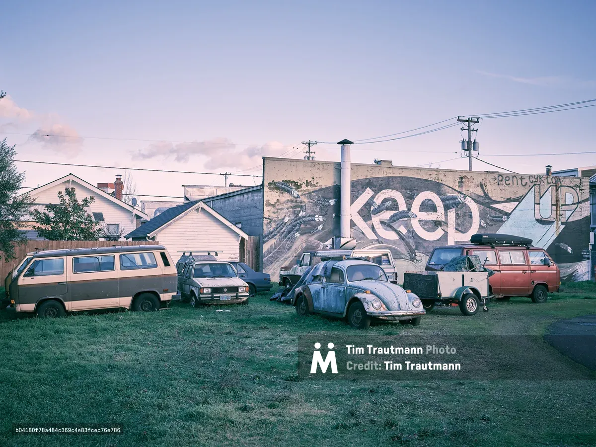 A collection of aging and worn vehicles, including two Volkswagen Vanagons, a rusted VW Beetle, and a red van with a roof rack, are parked on a grassy lot in Portland's Alberta Arts District. Behind them, a large weathered building displays a partial mural reading "Keep...Up," part of the "Keep Your Chin Up" street art piece, featuring painted feathers and organic forms in black, white, and blue. Residential homes, utility poles, and a soft purple-hued evening sky are visible in the background.