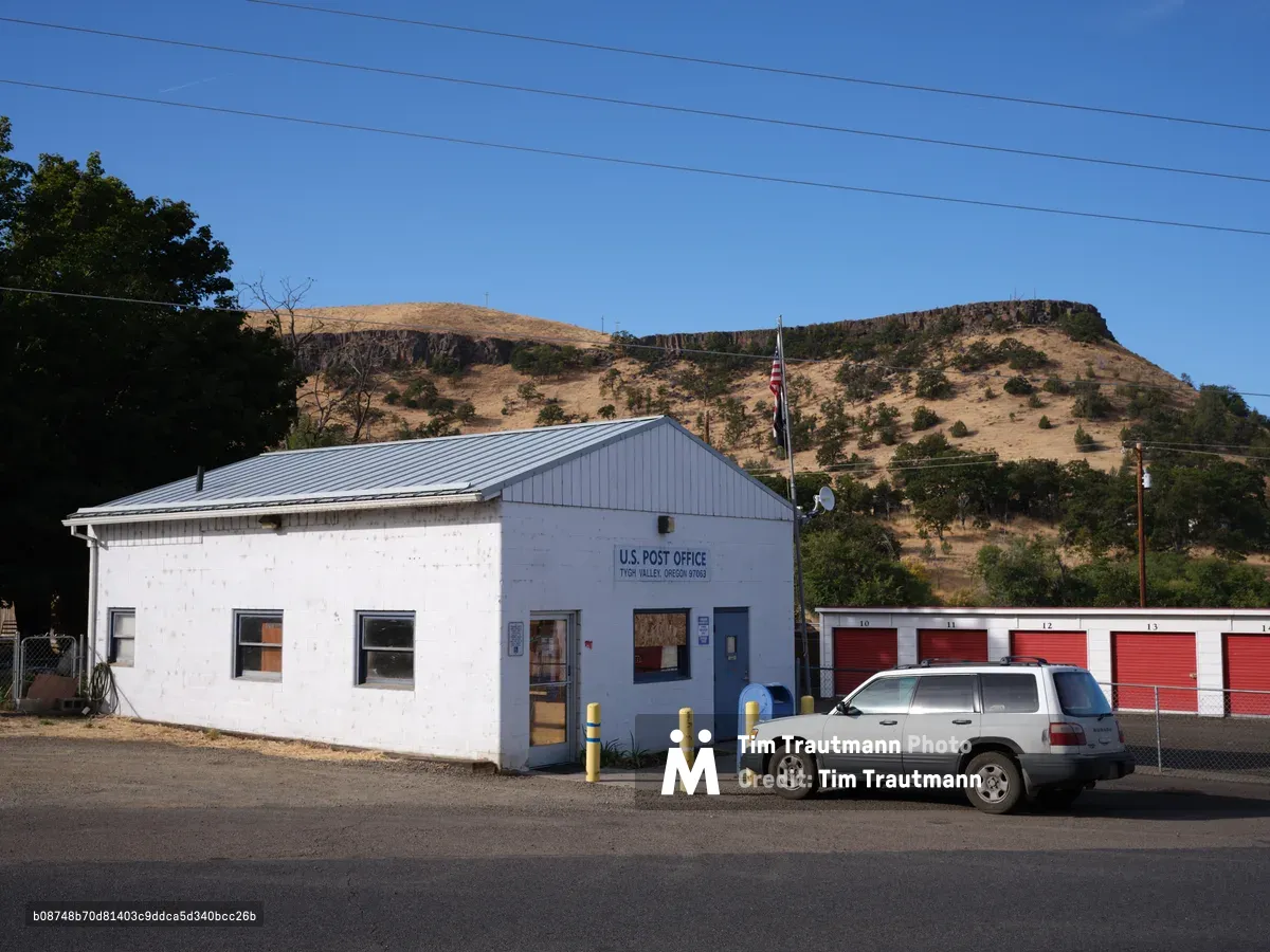 A modest white-painted U.S. Post Office building sits beneath the dramatic golden bluffs of Tygh Valley, Oregon, its metal roof gleaming under the crystalline blue sky. The humble single-story structure, flanked by red storage units and a weathered SUV, embodies the essential services that anchor rural American communities. Power lines streak across the azure expanse above sun-baked hills dotted with sparse vegetation, while shadows from mature trees provide relief from the high desert light. The scene captures the quiet dignity of small-town infrastructure against the raw beauty of the Columbia River Gorge region.