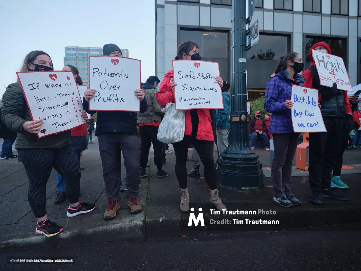 Healthcare workers gather on a grey Portland morning outside the Kaiser Permanente Tower in the Lloyd District, their handmade protest signs creating stark white rectangles against the building's glass facade. Masked nurses in winter jackets hold placards declaring "Patients Over Profits" and "Safe Staffing Saves Lives," their determination evident despite the overcast sky. The urban sidewalk scene captures the tension between corporate healthcare and frontline workers, with the tower's reflective windows looming behind the small but resolute group of demonstrators.