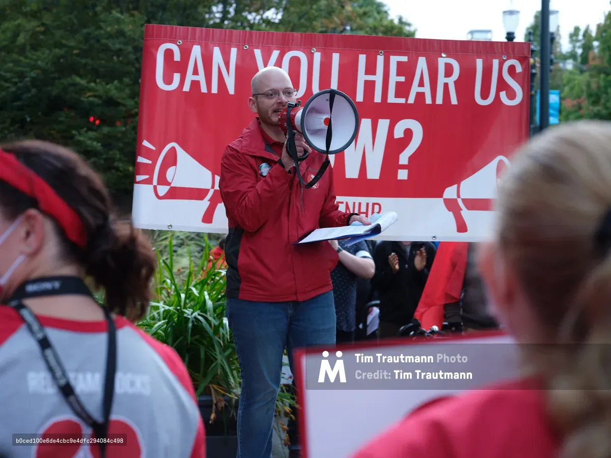 A bald man in a vibrant red jacket commands attention as he speaks through a megaphone at a Kaiser Permanente nurses' strike rally in Portland's Lloyd District. Behind him, a bold red and white banner poses the rhetorical question 'CAN YOU HEAR US NOW?' - its message emphasized by megaphone graphics. The speaker holds papers in one hand while addressing a crowd of supporters wearing matching red and white strike colors, creating a sea of solidarity that fills the foreground with purposeful energy.