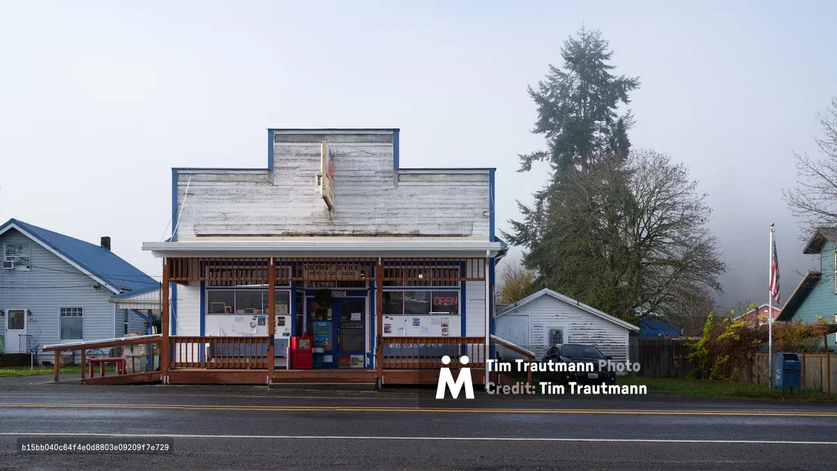 A weathered white post office building stands as the beating heart of rural Menlo, Washington, its false front facade bearing the scars of Pacific Northwest weather. The structure's wood-sided exterior and covered porch with simple railings evoke classic Americana, while an American flag and neon 'OPEN' sign signal its continued service to this small community. Towering evergreens frame the scene against a brooding gray sky, creating an atmosphere of quiet resilience along the empty two-lane road.