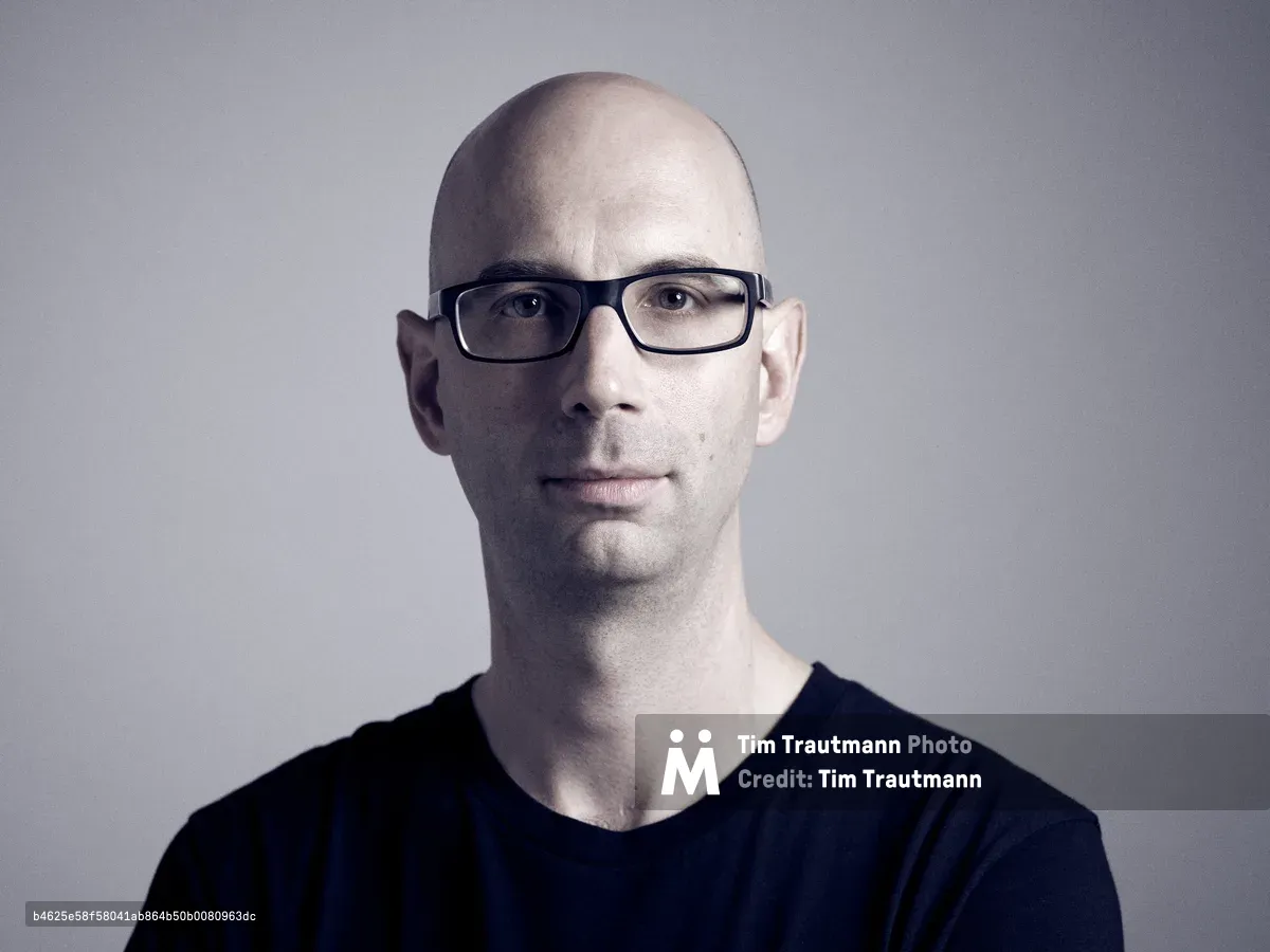 DJ and electronic music producer Jack Coleman sits for an intimate studio portrait against a refined gray backdrop in Portland's creative Central Eastside district. The controlled lighting creates subtle shadows that sculpt his bald head and black-framed glasses, while his dark v-neck shirt dissolves into shadow, focusing attention on his contemplative expression. The minimalist composition reflects the precision and artistry of his electronic music work with labels like Trapez and Translucent.