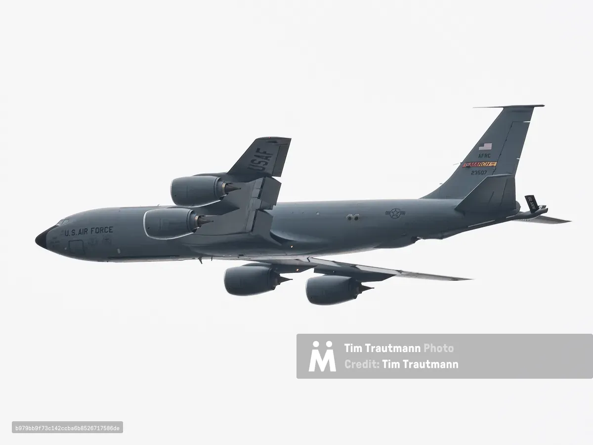 A U.S. Air Force Boeing KC-135 Stratotanker aerial refueling aircraft photographed in flight from below against a flat, overcast white sky. The aircraft is painted in overall grey and bears "U.S. AIR FORCE" lettering on the fuselage. "USAF" is stenciled on the vertical stabilizer, with "AFRC" and "MARCH" unit markings and the tail number 23507 visible near the tail. Four podded jet engines are mounted under the swept wings. The aircraft's boom refueling equipment is visible extending from the rear of the fuselage. A U.S. national insignia roundel appears on the fuselage. The tight telephoto framing isolates the aircraft cleanly against the grey-white sky.