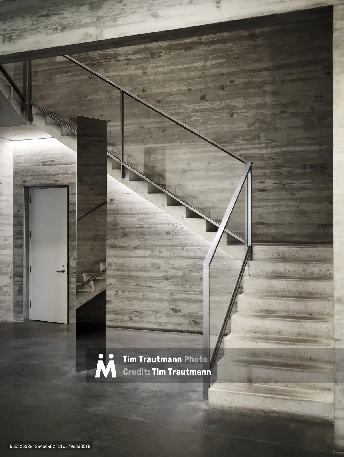A monumental concrete staircase ascends through raw architectural space, its board-formed surfaces bearing the ghostly imprints of wooden planks. Steel handrails trace geometric lines against weathered concrete walls, while muted natural light filters through the brutalist interior. The composition emphasizes the interplay between rough-hewn materiality and precise modern engineering, creating a cathedral-like atmosphere within this Chelsea arts district building.