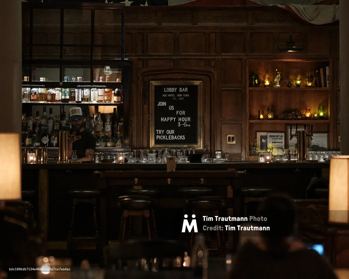 Amber light bathes the mahogany-paneled lobby bar at Manhattan's Ace Hotel, where a baseball-capped bartender works behind gleaming glassware. The vintage chalkboard advertises happy hour picklebacks while warm illumination from pendant lights creates intimate pools of light across the polished bar surface. Rich wood grain and carefully curated spirits bottles create an atmosphere of sophisticated urban refuge in the heart of NoMad.