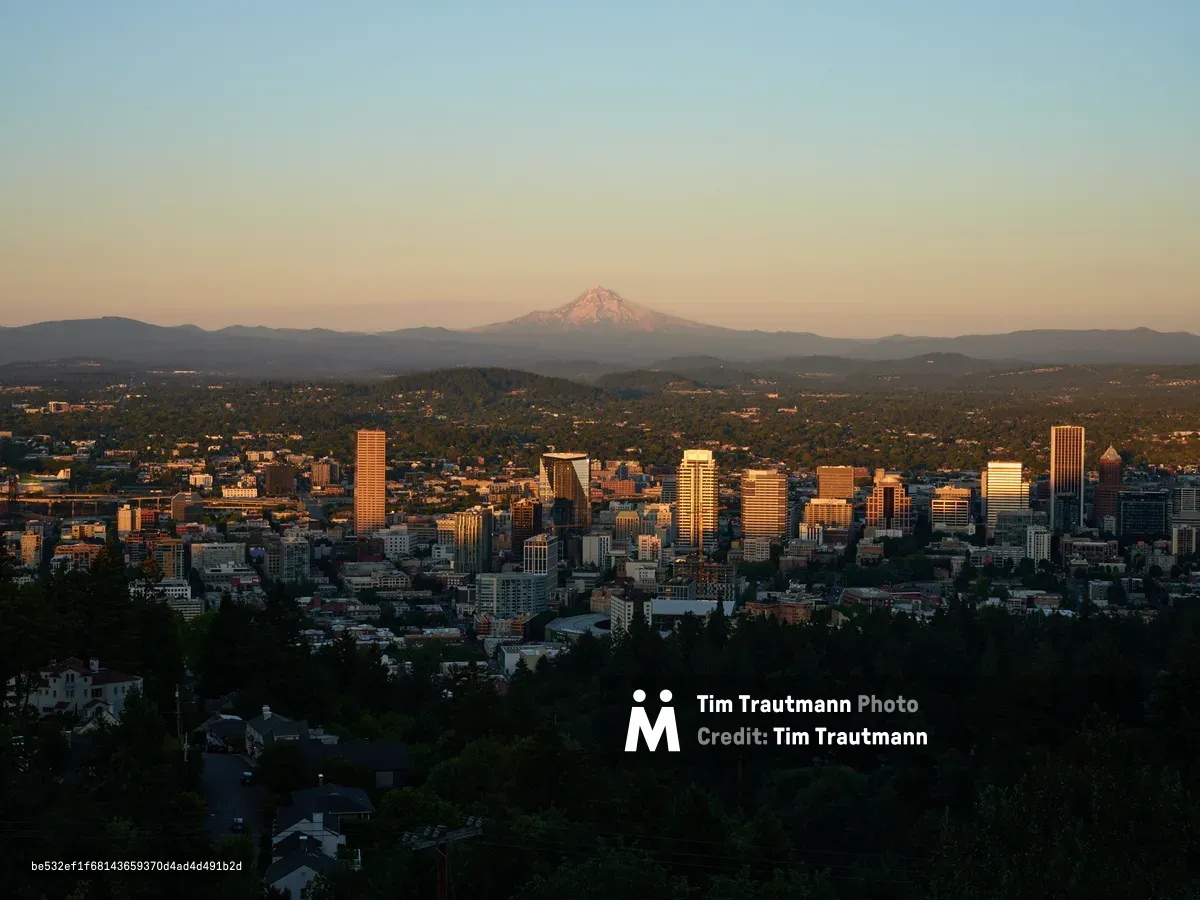 An elevated view of Portland, Oregon's downtown skyline at golden hour, with the snow-capped peak of Mount Hood visible in the distance beyond rolling hills. The city's high-rise buildings are illuminated by warm sunset light, while residential areas and dense forest cover the foreground and surrounding landscape.