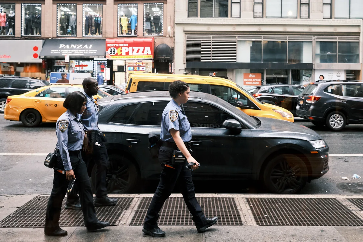 Three uniformed police officers stride purposefully along 7th Avenue in Manhattan's Chelsea District, their light blue shirts and dark equipment belts catching the filtered daylight. Behind them, yellow taxi cabs and a sleek black sedan are locked in typical New York traffic, while storefronts including a pizza joint and clothing displays create the quintessential urban backdrop. The scene captures the rhythm of city law enforcement moving through the congested arteries of midtown Manhattan, where pedestrian and vehicular worlds intersect in perpetual motion.