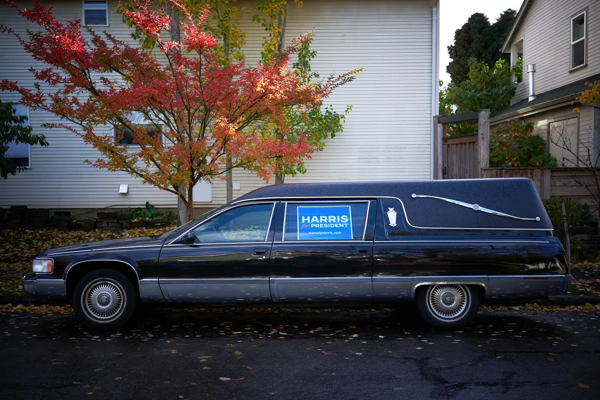A black funeral hearse parked on a residential street in Portland, Oregon displays a blue Harris for President campaign sign in its window. The vehicle sits beneath autumn trees with vibrant red foliage, with white wooden houses visible in the background.