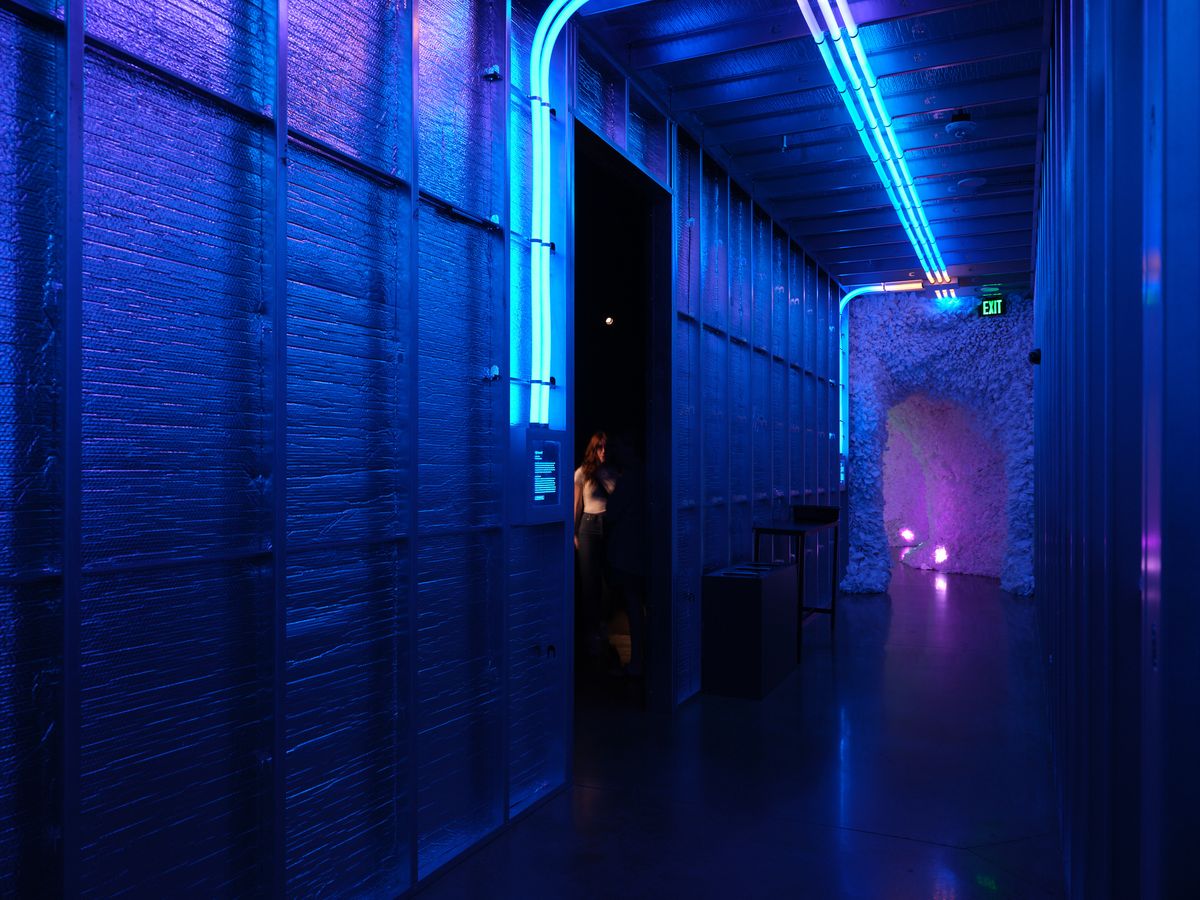 A dramatic blue-lit corridor in Hopscotch, Portland, featuring exposed brick walls, neon lighting, and a person silhouetted in a doorway. The industrial space combines modern lighting design with historic architecture.