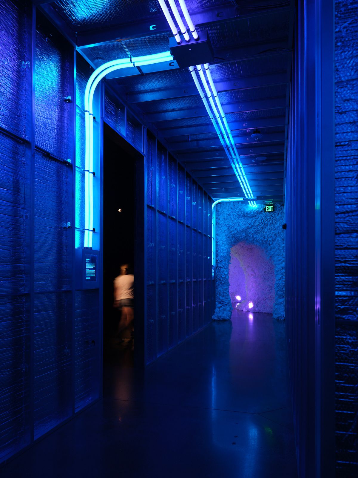 A atmospheric hallway with dramatic blue neon lighting illuminating textured walls and exposed ceiling elements, with a blurred figure walking through a doorway. The modern industrial interior features continuous LED strip lighting along the ceiling and walls creating a futuristic ambiance.