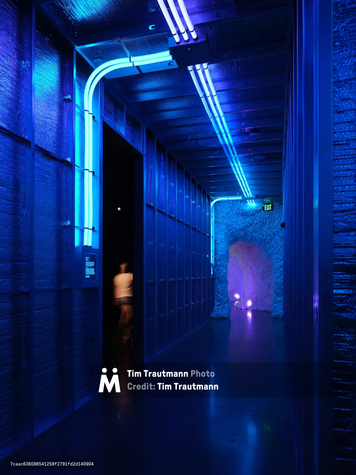 A atmospheric hallway with dramatic blue neon lighting illuminating textured walls and exposed ceiling elements, with a blurred figure walking through a doorway. The modern industrial interior features continuous LED strip lighting along the ceiling and walls creating a futuristic ambiance.