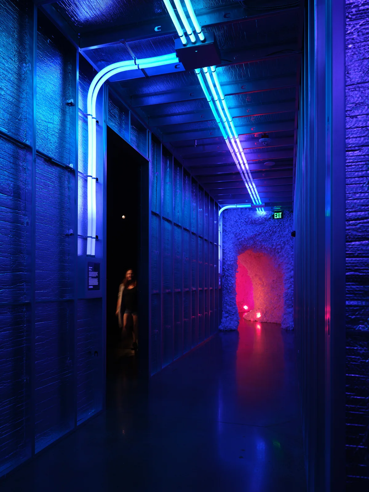 A dramatic industrial hallway illuminated by bright blue and cyan neon lighting, with exposed brick walls and ceiling beams creating a cyberpunk atmosphere. A silhouetted figure stands in a doorway while warm red light glows from an alcove at the corridor's end.