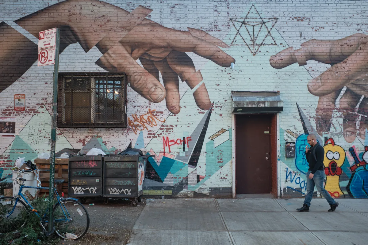 A lone figure in a black leather jacket strides past a vibrant mural on weathered brick walls in Williamsburg, Brooklyn. The street art features massive reaching hands, geometric patterns, and colorful character illustrations that transform the urban facade into a canvas of contemporary expression. A blue bicycle rests against dumpsters in the foreground, while peeling paint and layered graffiti tags speak to the neighborhood's authentic street culture. The overcast lighting creates a moody atmosphere that captures the raw creative energy of Brooklyn's artistic landscape.