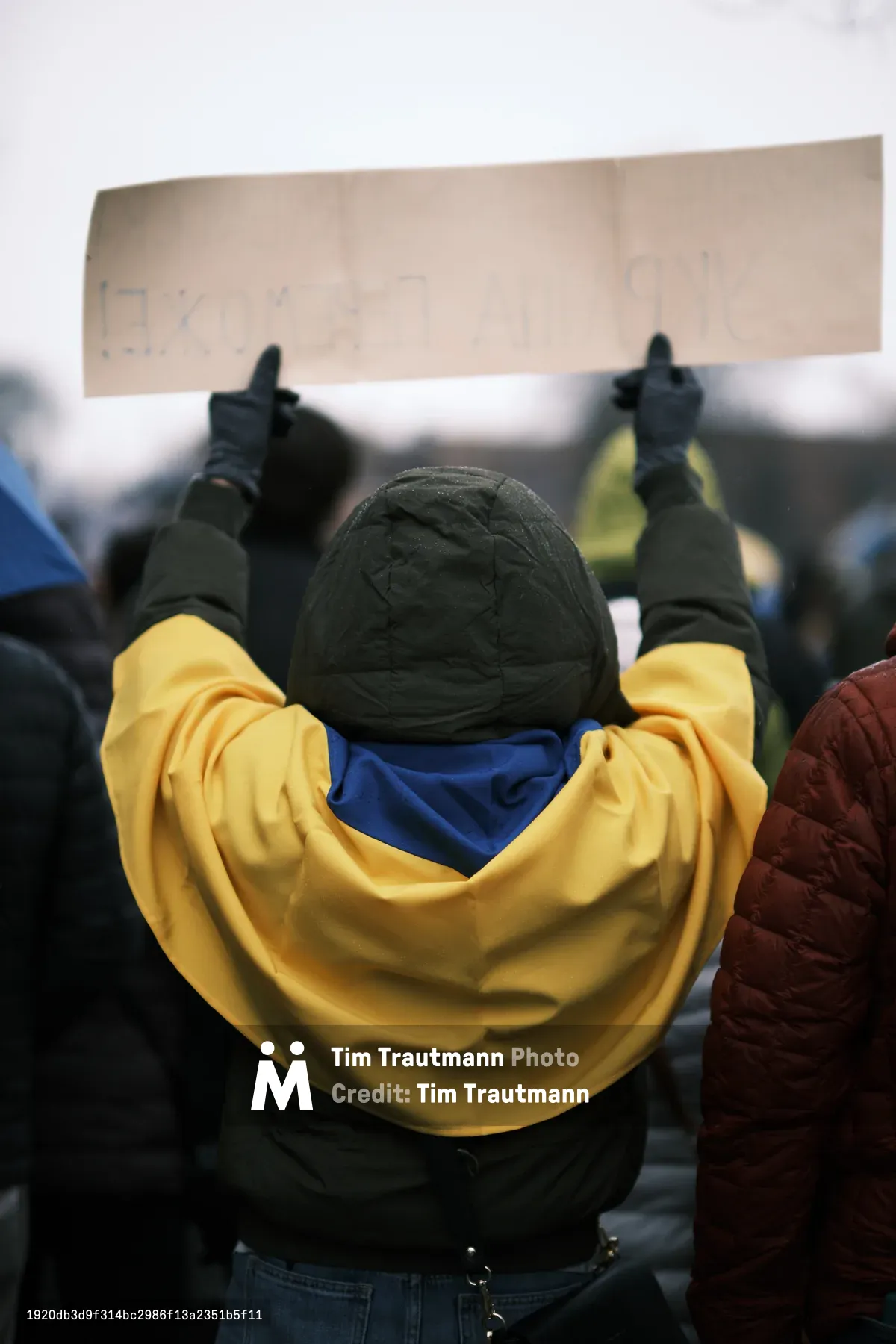 A lone protester stands resolute outside Portland's Revolution Hall, their face obscured by a quilted beanie and wrapped in the symbolic blue and yellow of Ukraine's flag. Dark winter gloves grip a handwritten sign overhead, while the golden fabric pools around their shoulders like a protective embrace against both the February chill and the weight of distant conflict. The shallow depth of field dissolves the crowd into a soft blur, isolating this singular act of solidarity in the days before Russian forces would cross into Ukrainian territory.