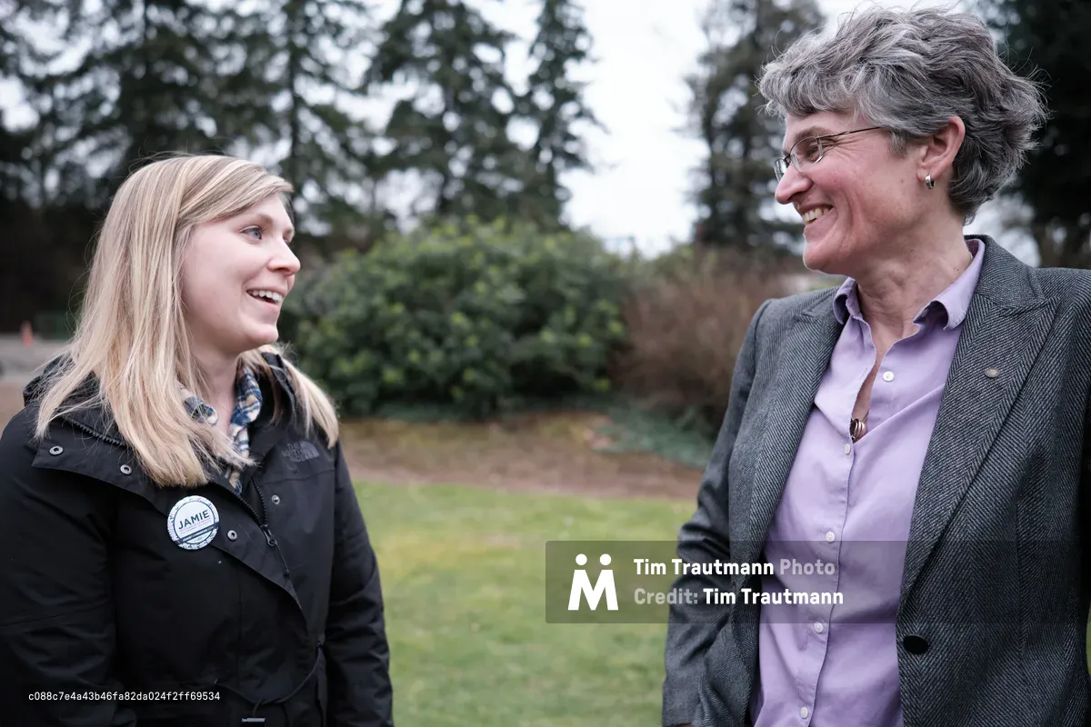 Two women stand facing each other in an outdoor park setting in Oregon City, Oregon, engaged in friendly conversation and smiling. The younger blonde woman on the left wears a dark jacket with a campaign button, while the older woman on the right wears glasses, a gray blazer, and purple shirt.