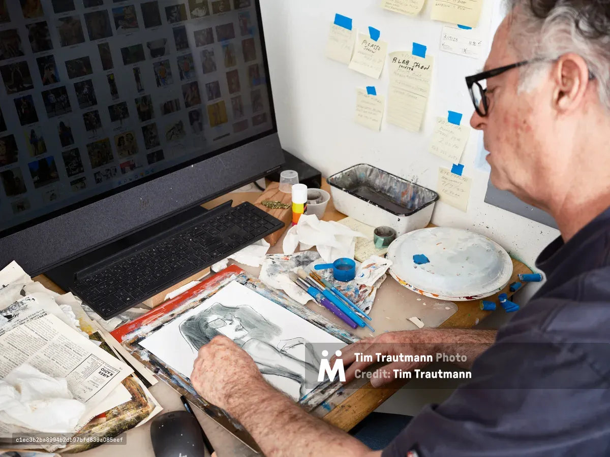 An older man with glasses sits at a cluttered desk in his Portland art studio, working on a portrait drawing while surrounded by art supplies, reference images on his laptop screen, and notes taped to the wall.